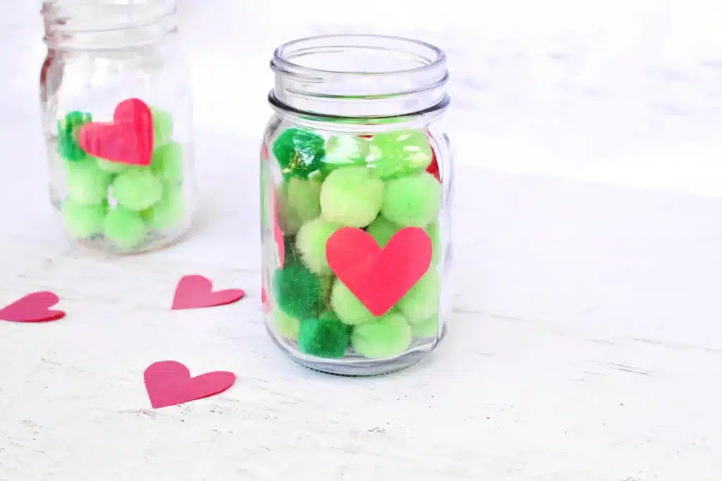 Grinch Heart Jar Challenge - Cultivating Brilliant Minds a mason jar filled with green pom poms and a paper heart taped on