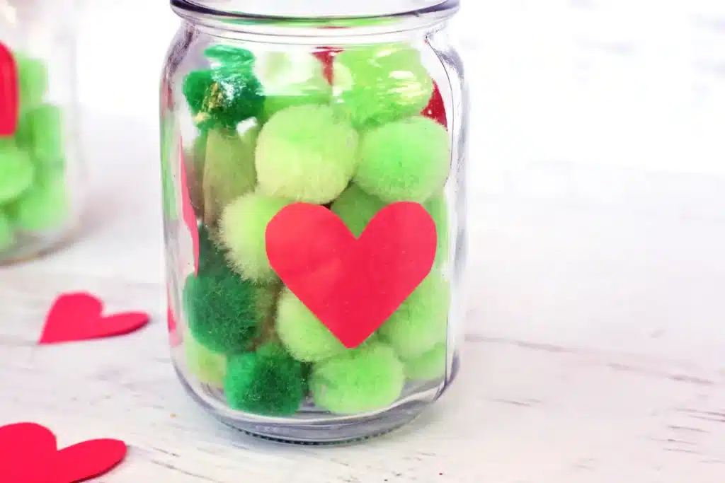 Grinch Heart Jar Challenge - Cultivating Brilliant Minds a mason jar filled with green pom poms and a paper heart taped on