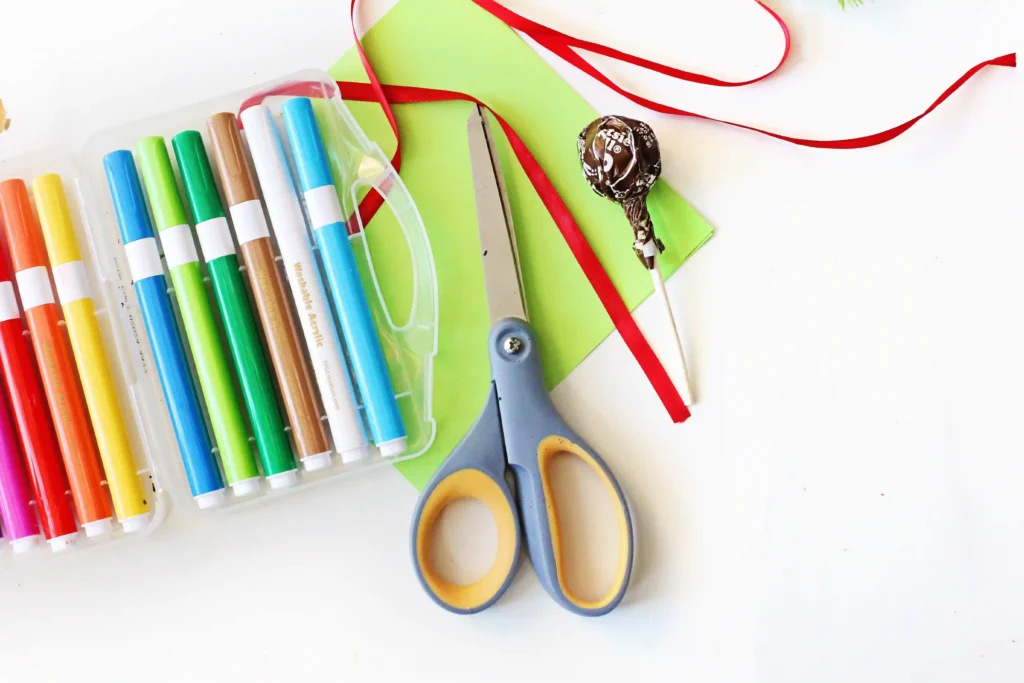 Grinch Lollipops - Cultivating Brilliant Minds green tissue paper, makers, red ribbon, scissors, and a lollipop