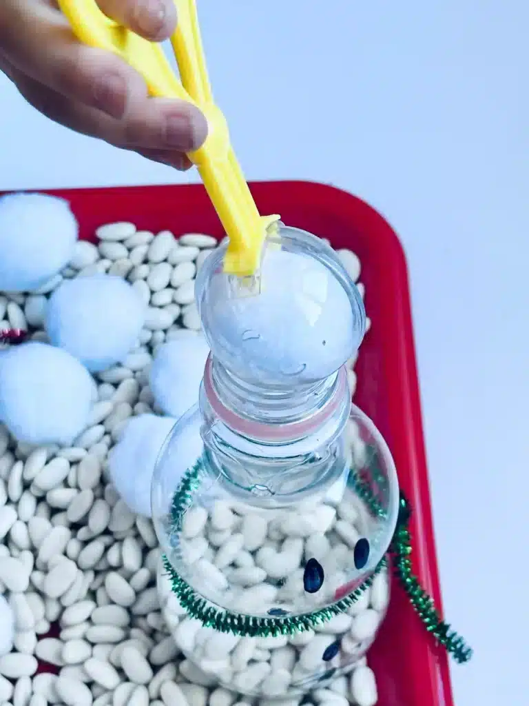 Snowman Sensory Bin - Cultivating Brilliant Minds a kid scooping a cotton ball and dropping it in a plastic bottle