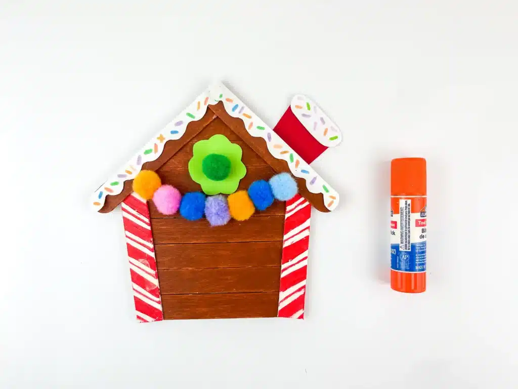 Gingerbread House Craft - Cultivating Brilliant Minds a gingerbread house made of popsicle sticks painted brown and pom poms