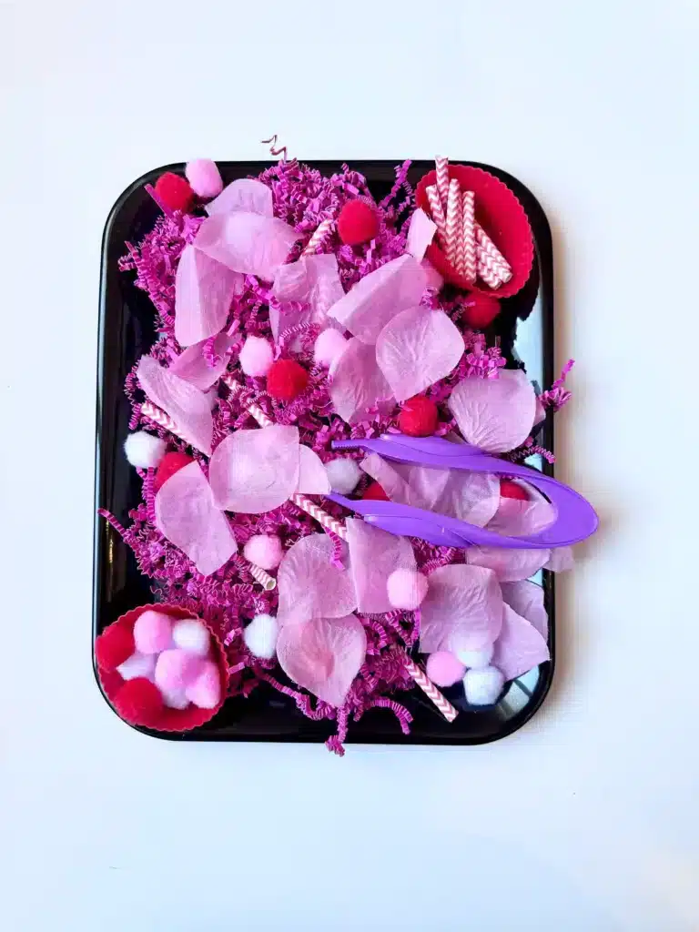 a valentine's day sensory bin with crinkle paper. rose petals, pom poms, and straws