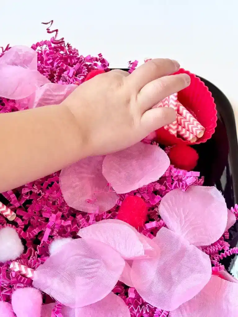 Valentine's Day Sensory Bin - Cultivating Brilliant Minds a kid picking up a straw in the valentine's day sensory bin with crinkle paper. rose petals, pom poms, and straws