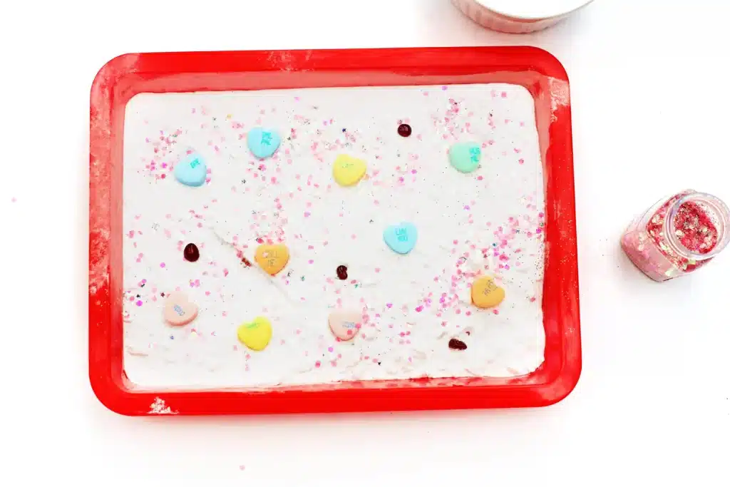 Valentine's Day Fizzy Candy Hearts Experiment - Cultivating Brilliant Minds a red tray with a baking soda layer on the bottom and candy hearts, glitter, and red food dye
