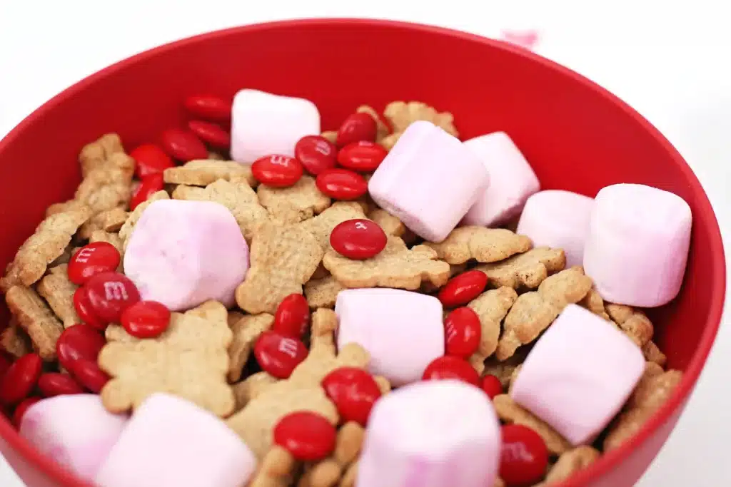 Valentine's Day Cupid Snack Mix - Cultivating Brilliant Minds a red bowl with snack mix inside - pink marshmallows, red m&ms and teddy grahams