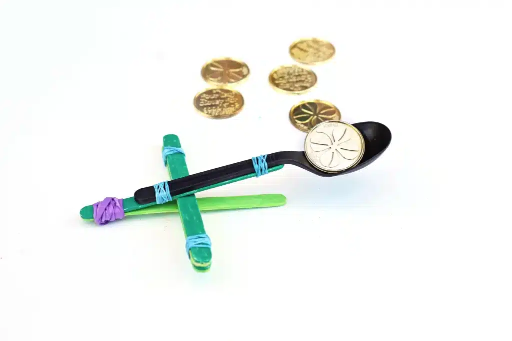 St. Patrick's Day Gold Coin Catapults - Cultivating Brilliant Minds a spoon attached to popsicle sticks with rubber bands to make a catapault