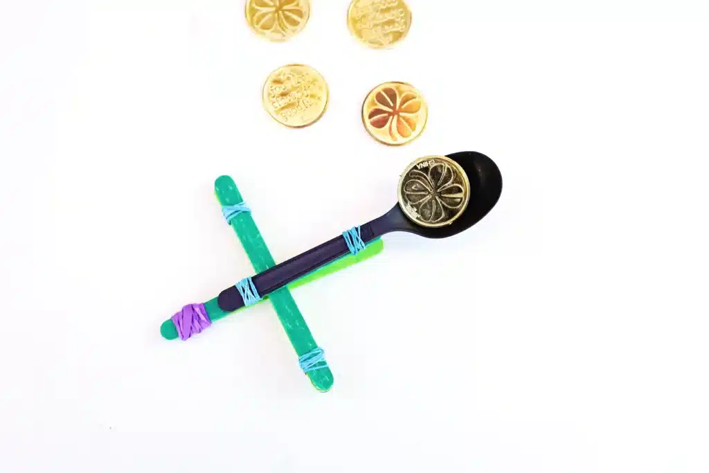 St. Patrick's Day Gold Coin Catapults - Cultivating Brilliant Minds a spoon attached to popsicle sticks with rubber bands to make a catapault