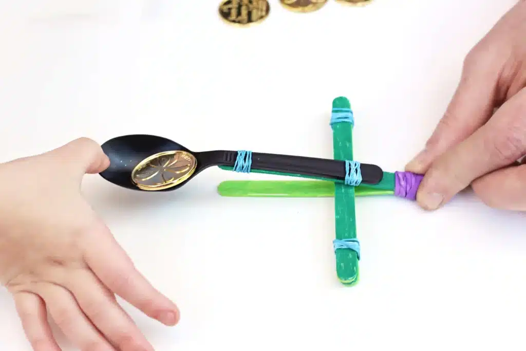 St. Patrick's Day Gold Coin Catapults - Cultivating Brilliant Minds a spoon attached to popsicle sticks with rubber bands to make a catapult