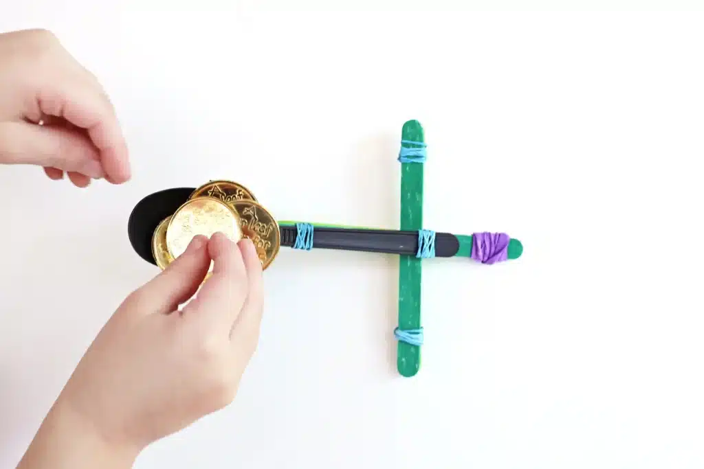 St. Patrick's Day Gold Coin Catapults - Cultivating Brilliant Minds a spoon attached to popsicle sticks with rubber bands to make a catapault