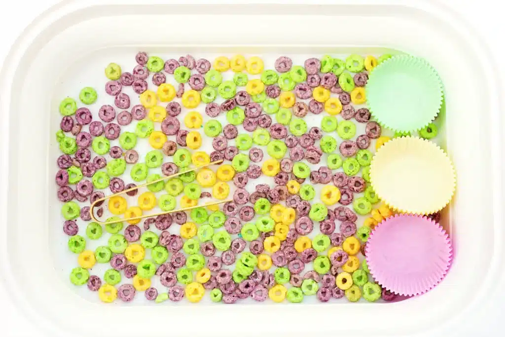 Mardi Gras Color Sorting Sensory Bin - Cultivating Brilliant Minds a bin filled with green, yellow, and purple froot loops, tongs, and green, yellow, and purple muffin tin liners
