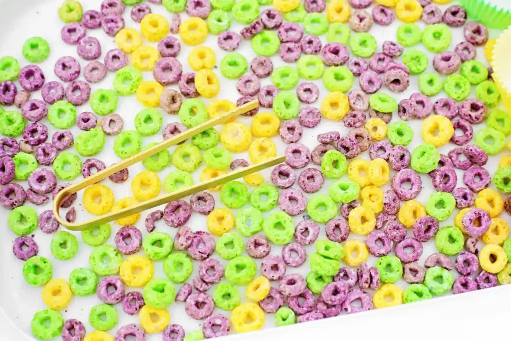 Mardi Gras Color Sorting Sensory Bin - Cultivating Brilliant Minds green, purple, and yellow fruit loops in a bin with tongs