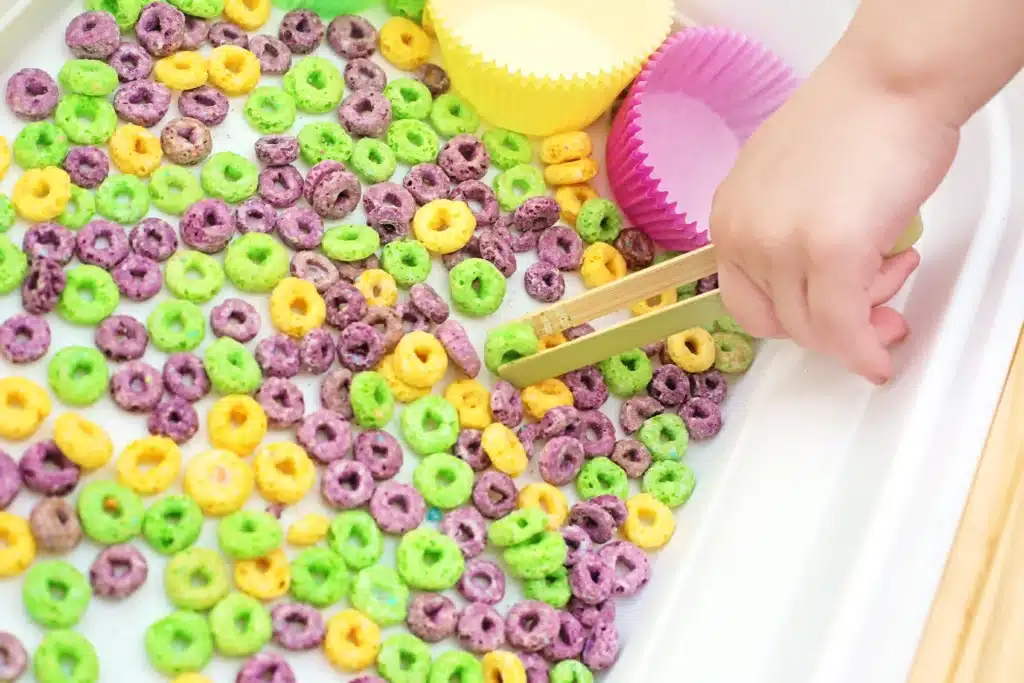 Mardi Gras Color Sorting Sensory Bin - Cultivating Brilliant Minds a kid using tongs to pick up froot loops