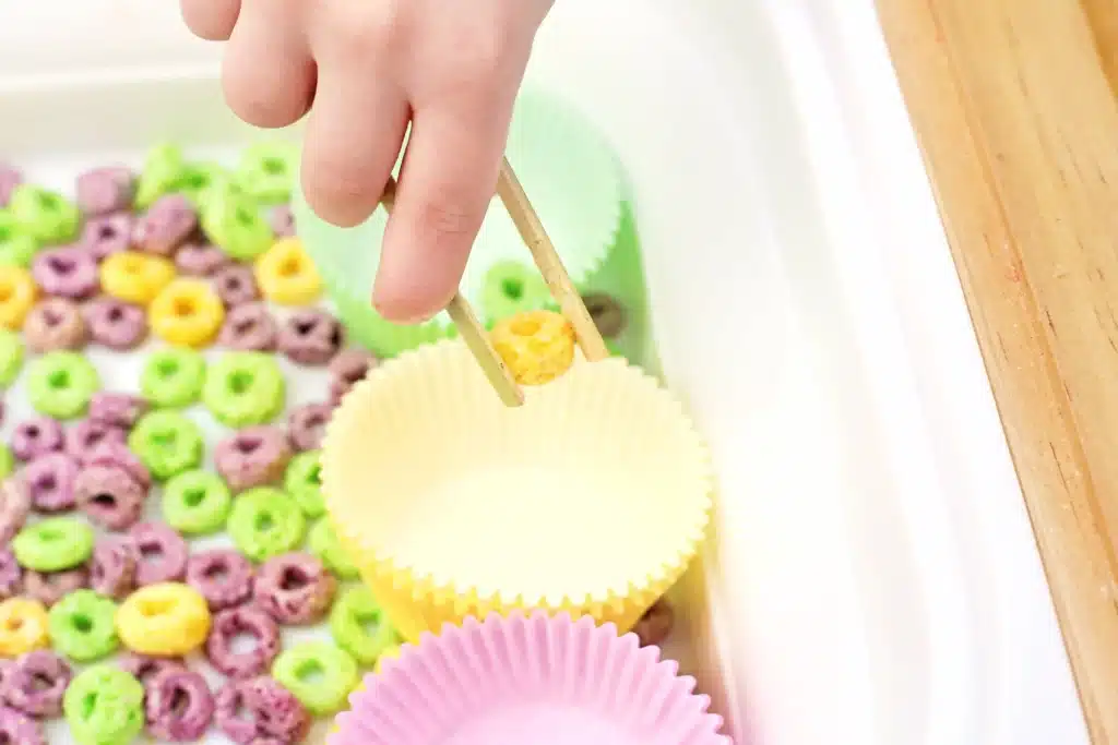 Mardi Gras Color Sorting Sensory Bin - Cultivating Brilliant Minds a kid using tongs to put a yellow froot loop in a yellow muffin tin liner