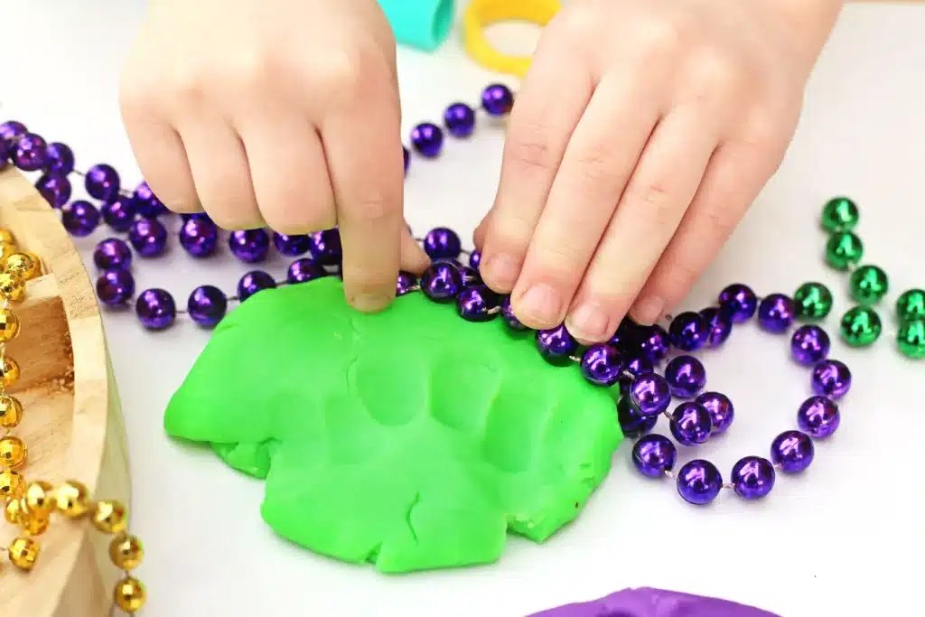 Mardi Gras Playdough Tray - Cultivating Brilliant Minds a kid pushing a purple beaded necklace into green playdough