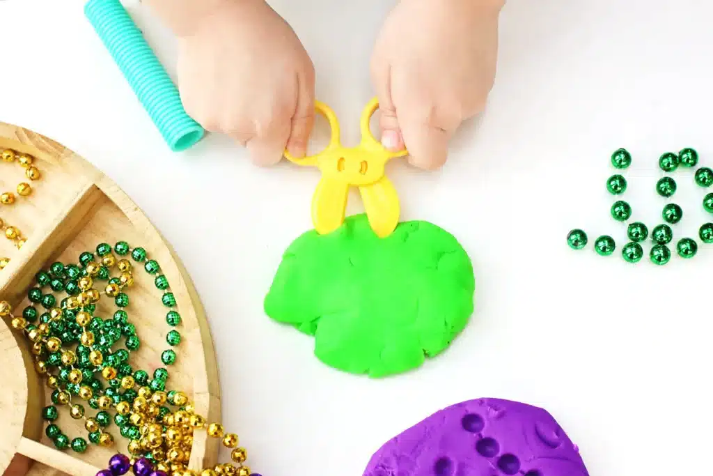 Mardi Gras Playdough Tray - Cultivating Brilliant Minds a kid using plastic scissors to cut green playdough