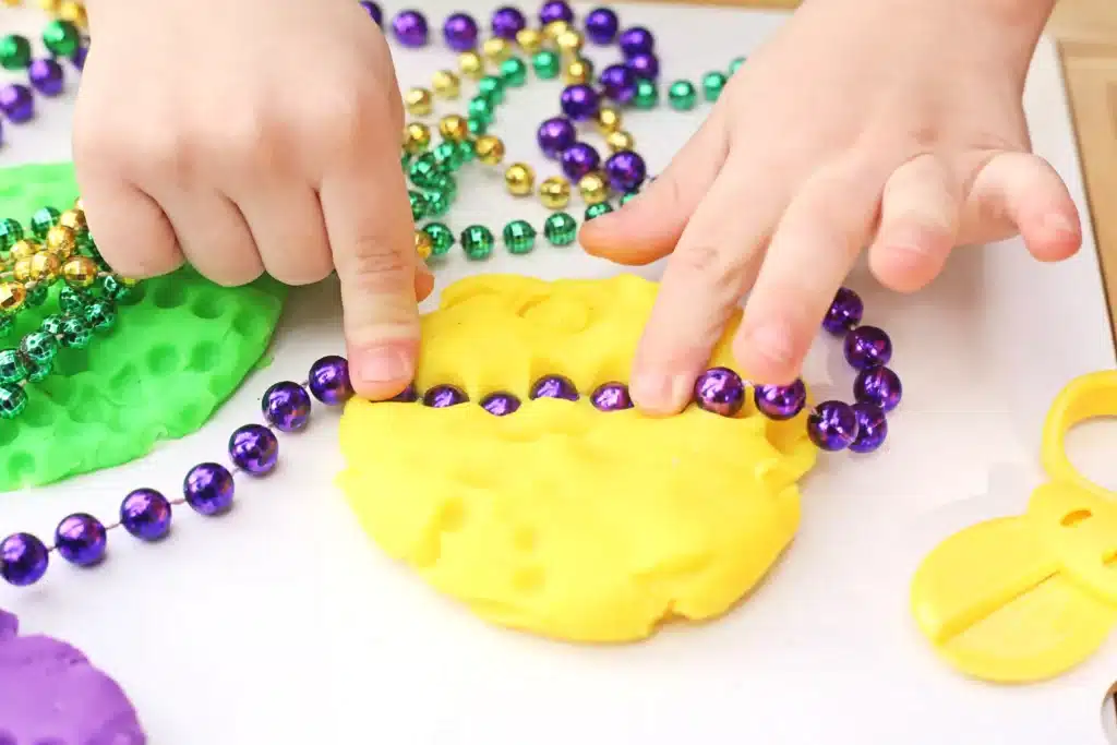 Mardi Gras Playdough Tray - Cultivating Brilliant Minds a kid pushing a purple beaded necklace into yellow playdough
