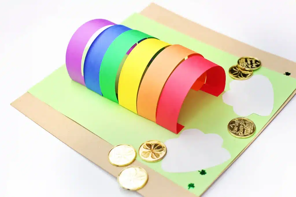 DIY Paper Leprechaun Trap STEM Craft - Cultivating Brilliant Minds colored construction paper glued to a piece of green paper. The colored construction paper is glued as an arch to catch a leprechaun. There are gold coins around it.