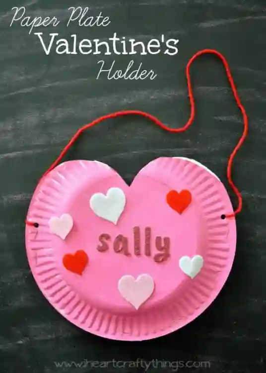 Heart-Themed Crafts For Kids This Valentine's Day - Cultivating Brilliant Minds a paper plate cut into a heart, painted pink and adorned with heart stickers, letter stickers that spell "sally" and red string.