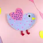 Lovebird Paper Plate Craft for Valentine's Day - Cultivating Brilliant Minds a paper plate cut and colored to look like a lovebird. It is painted purple with pink hearts and has a pink heart with purple hearts drawn all over it for a wing.