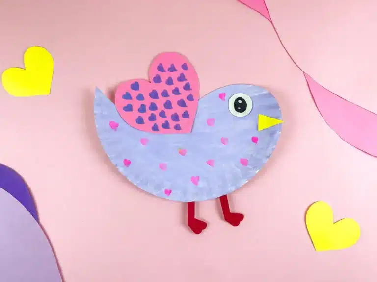 a paper plate cut and colored to look like a lovebird. It is painted purple with pink hearts and has a pink heart with purple hearts drawn all over it for a wing.