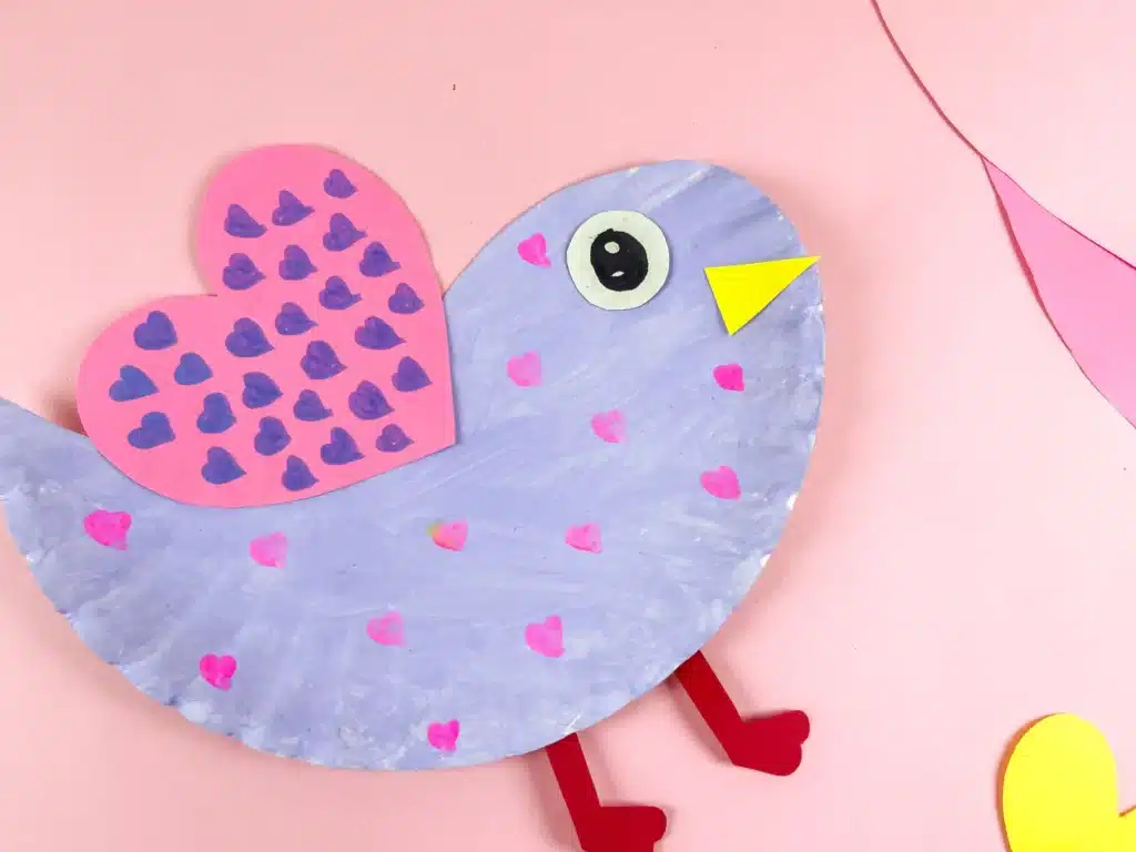 Lovebird Paper Plate Craft for Valentine's Day - Cultivating Brilliant Minds a lovebird paper plate craft that uses a paper plate cut and colored to look like a lovebird. It is painted purple with pink hearts and has a pink heart with purple hearts drawn all over it for a wing.