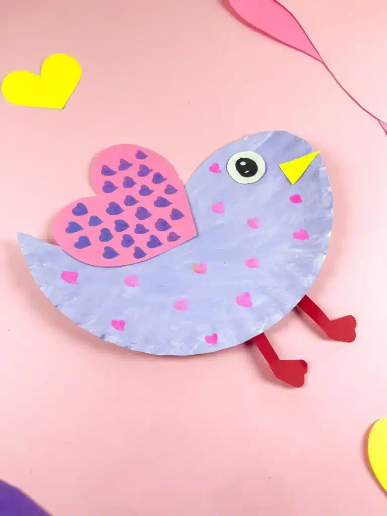Lovebird Paper Plate Craft for Valentine's Day - Cultivating Brilliant Minds a paper plate cut and colored to look like a lovebird. It is painted purple with pink hearts and has a pink heart with purple hearts drawn all over it for a wing.