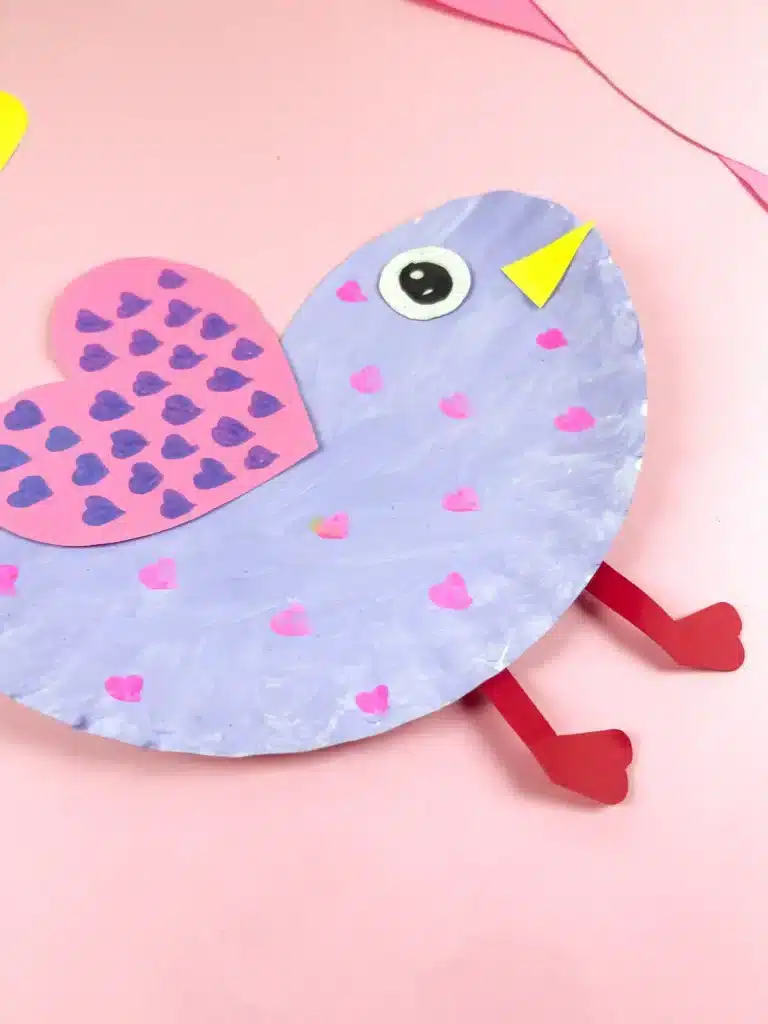 Lovebird Paper Plate Craft for Valentine's Day - Cultivating Brilliant Minds a paper plate cut and colored to look like a lovebird. It is painted purple with pink hearts and has a pink heart with purple hearts drawn all over it for a wing.
