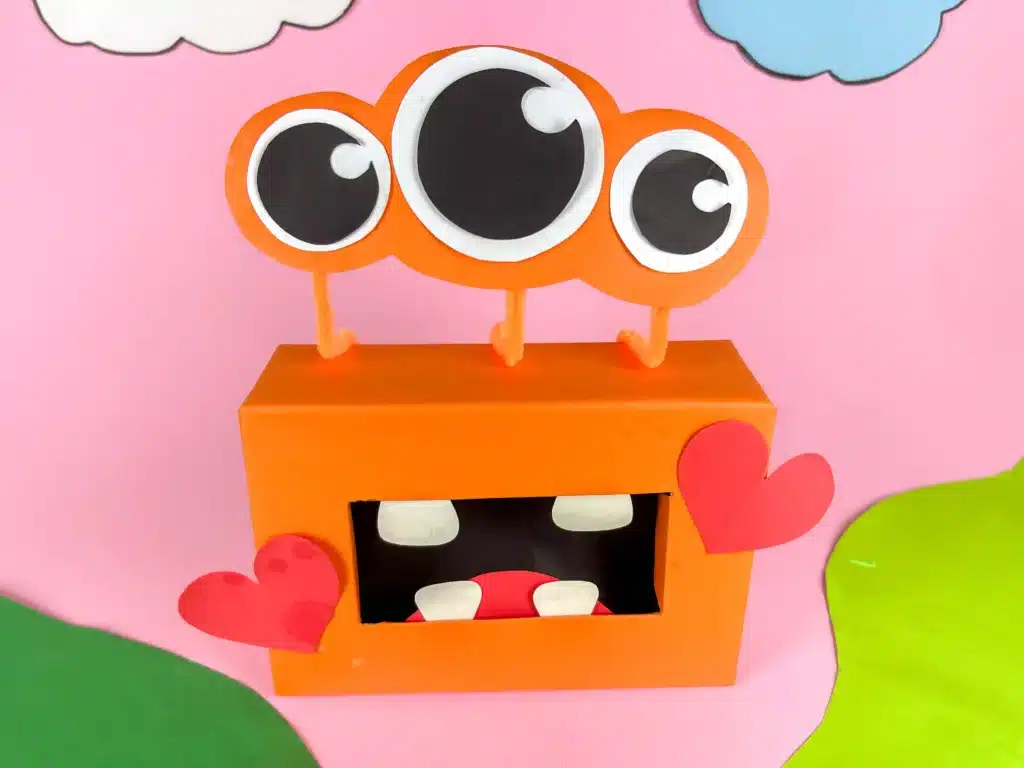 Monster Valentine Box - Cultivating Brilliant Minds a monster valentine box that has a box covered in orange paper, construction paper mouth and teeth and construction paper eyeballs attached using pipe cleaners