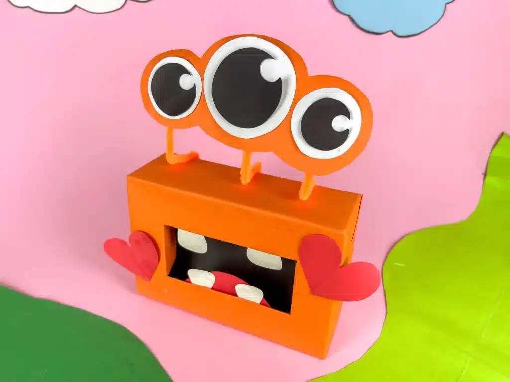 Monster Valentine Box - Cultivating Brilliant Minds a monster valentine box that has a box covered in orange paper, construction paper mouth and teeth and construction paper eyeballs attached using pipe cleaners
