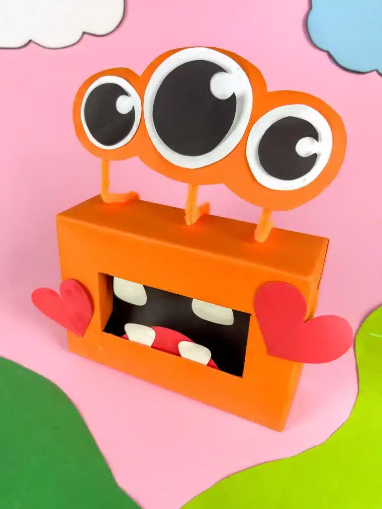Monster Valentine Box - Cultivating Brilliant Minds a monster valentine box that has a box covered in orange paper, construction paper mouth and teeth and construction paper eyeballs attached using pipe cleaners