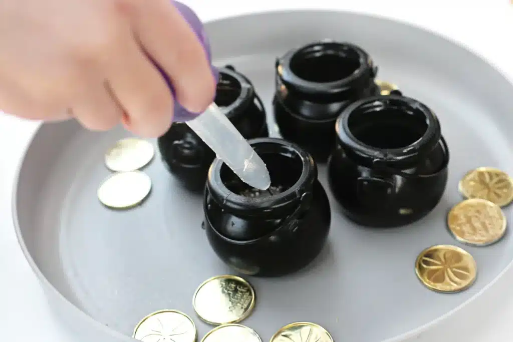 Fizzy Pot of Gold Experiment - Cultivating Brilliant Minds a kid using a pipette to add vinegar to a black cauldron of baking soda and gold gitter