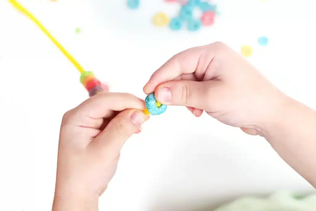 Froot Loops Rainbow Snack - Cultivating Brilliant Minds a child threading a froot loop onto a pipe cleaner