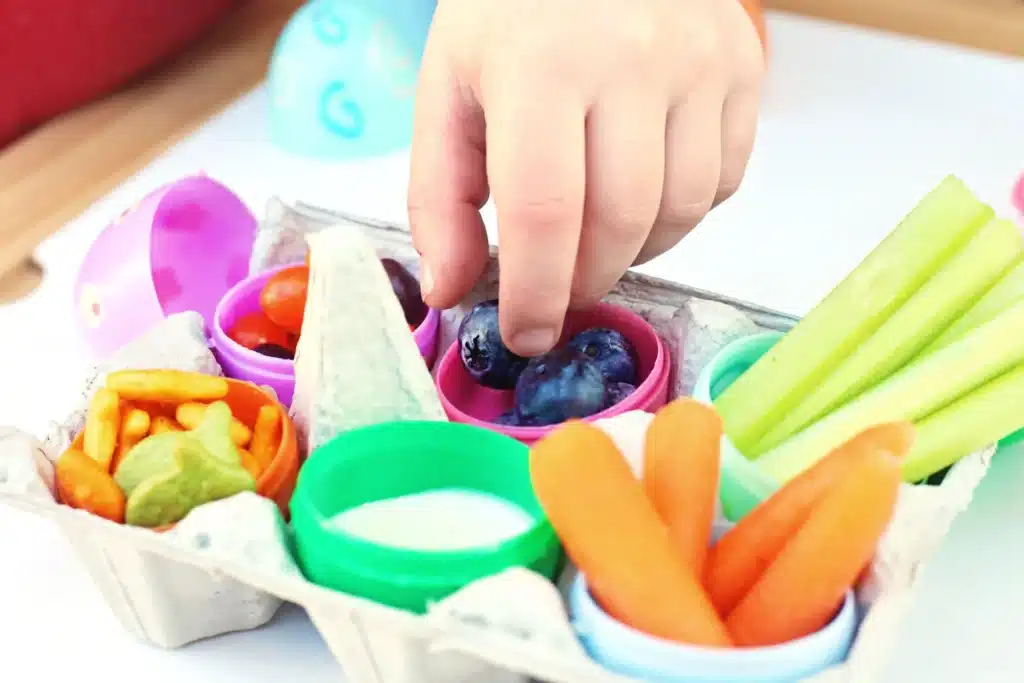 Easter Egg Snack Tray - Cultivating Brilliant Minds a kid grabbing a blueberry from an easter egg snack tray with an egg carton with plastic eggs inside and the eggs have jelly beans, blueberries, celery, carrots, goldfish, and ranch dressing inside