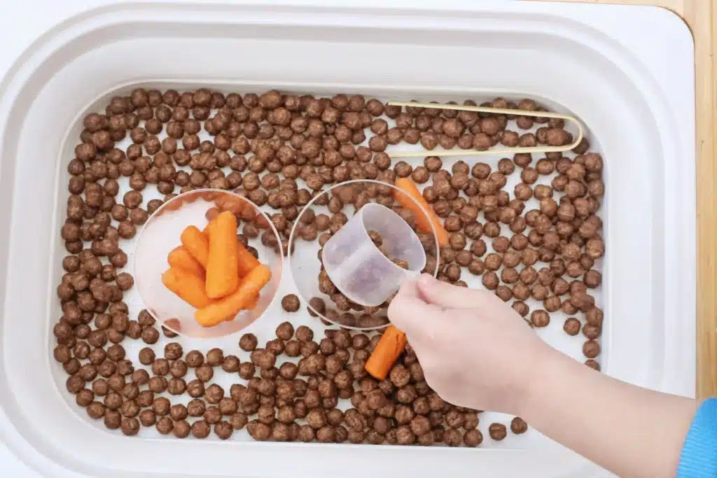 Edible Carrot Patch Sensory Bin For Spring - Cultivating Brilliant Minds a kid dumping cocoa puffs from a measuring cup