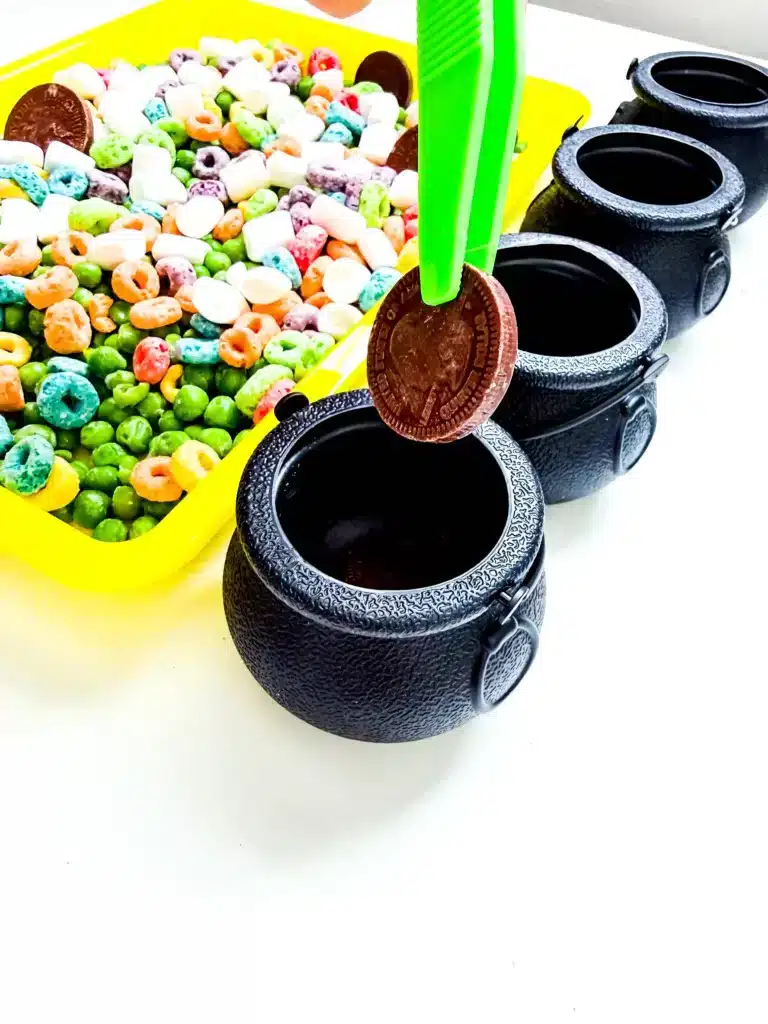 Edible St. Patrick's Day Sensory Bin - Cultivating Brilliant Minds tweezers holding a chocolate coin over a cauldron