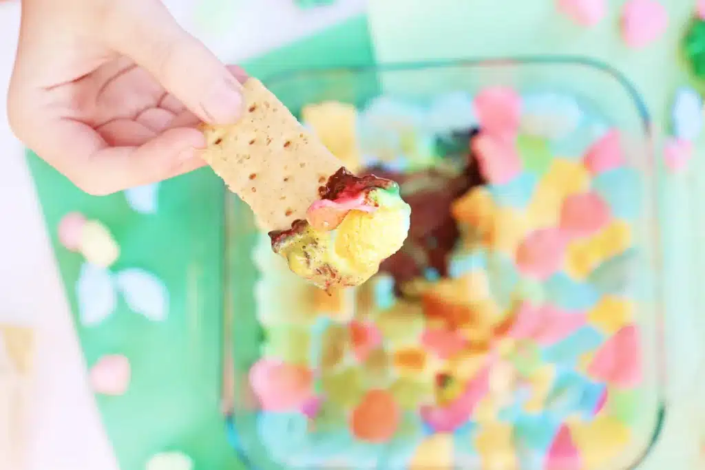 Lucky Charms S'mores Dip - Cultivating Brilliant Minds a graham cracker dipped into a lucky charms s'mores dip that has melted chocolate a lucky charm marshmallows