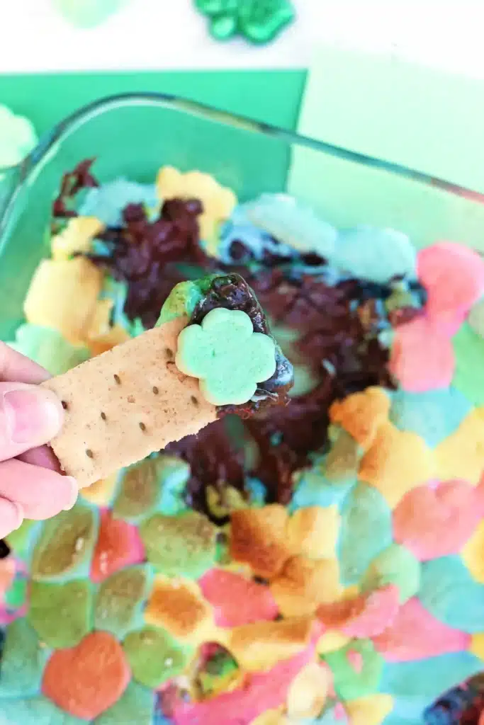 Lucky Charms S'mores Dip - Cultivating Brilliant Minds a graham cracker dipped into a lucky charms s'mores dip that has melted chocolate a lucky charm marshmallows