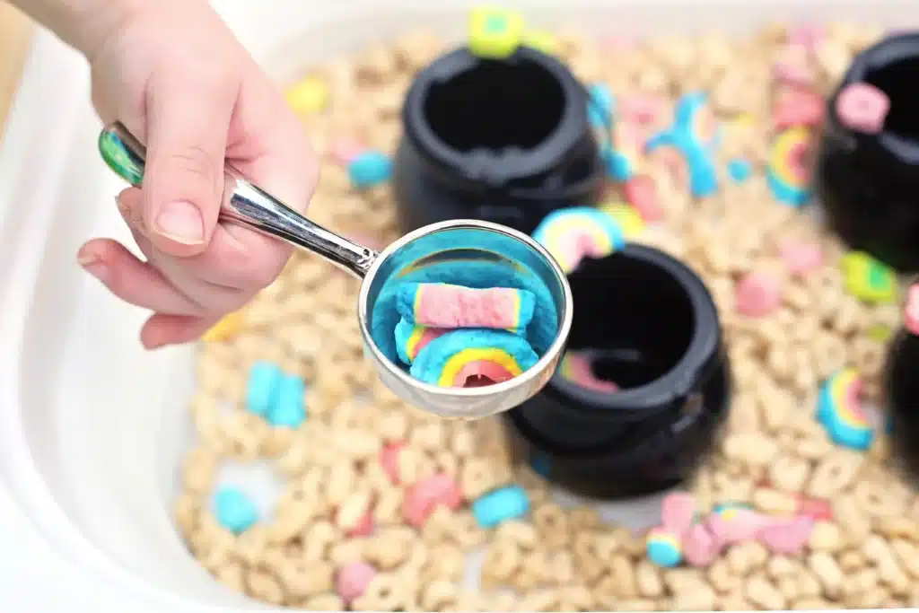 Lucky Charms Sensory Bin - Cultivating Brilliant Minds a kid with a measuring cup of rainbow marshmallows from lucky charms cereal
