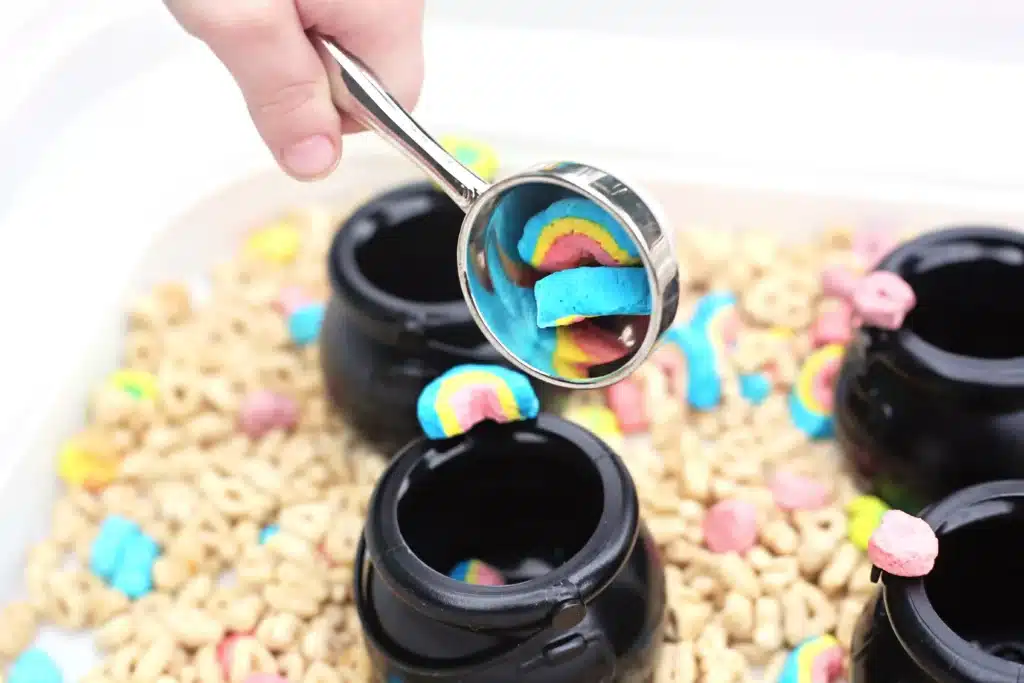 Lucky Charms Sensory Bin - Cultivating Brilliant Minds a kid with a measuring cup full of rainbow marshmallows from lucky charms cereal dumping them into a black cauldron pot