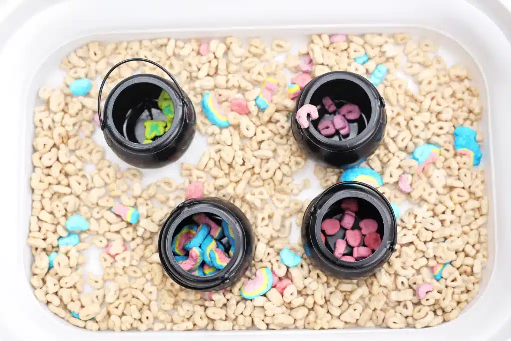 Lucky Charms Sensory Bin - Cultivating Brilliant Minds a sensory bin that uses lucky charms cereal, a scoop, and mini black cauldron pots