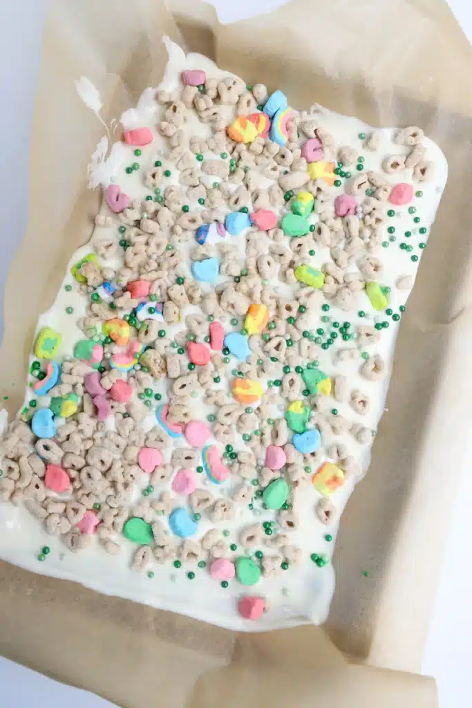 LuckyCharmsBarkInProcess-11 - Cultivating Brilliant Minds melted white chocolate spread on parchment paper with lucky charms cereal and green sprinkles on top
