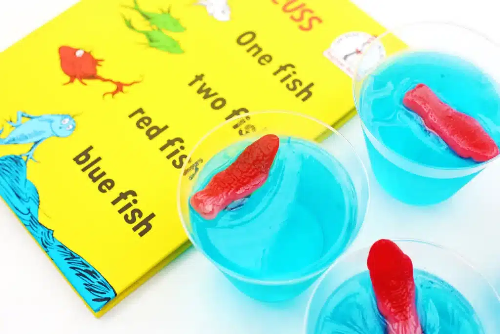 Dr. Seuss One Fish Two Fish Jello Cups - Cultivating Brilliant Minds blue jello cups with red swedish fish on top next to Dr. Seuss's One fish, two fish, red fish, blue fish book
