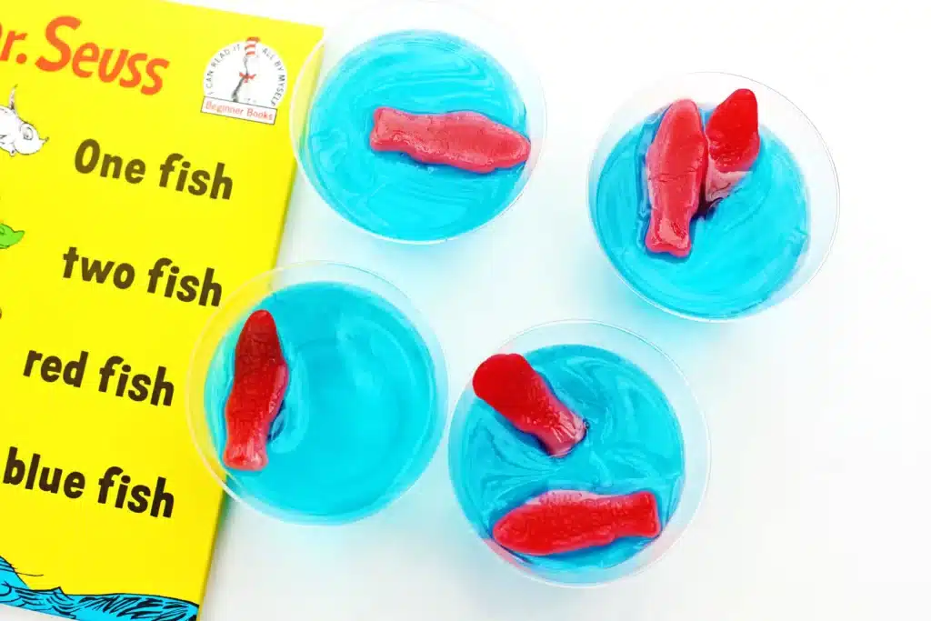 Dr. Seuss One Fish Two Fish Jello Cups - Cultivating Brilliant Minds blue jello cups with red swedish fish on top next to Dr. Seuss's One fish, two fish, red fish, blue fish book