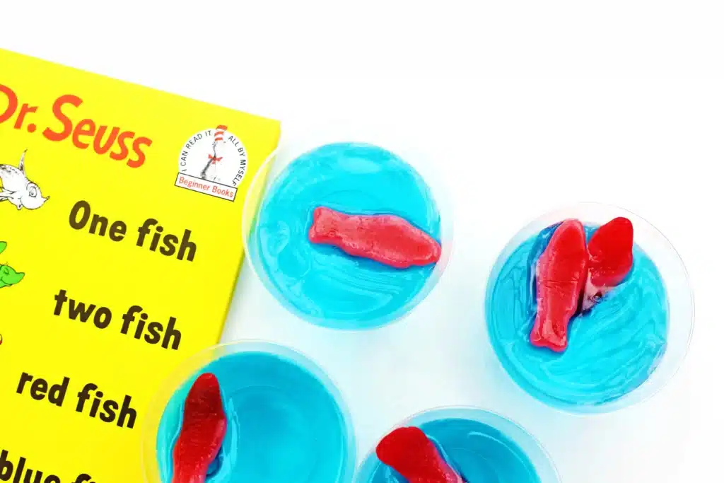 Dr. Seuss One Fish Two Fish Jello Cups - Cultivating Brilliant Minds blue jello cups with red swedish fish on top next to Dr. Seuss's One fish, two fish, red fish, blue fish book