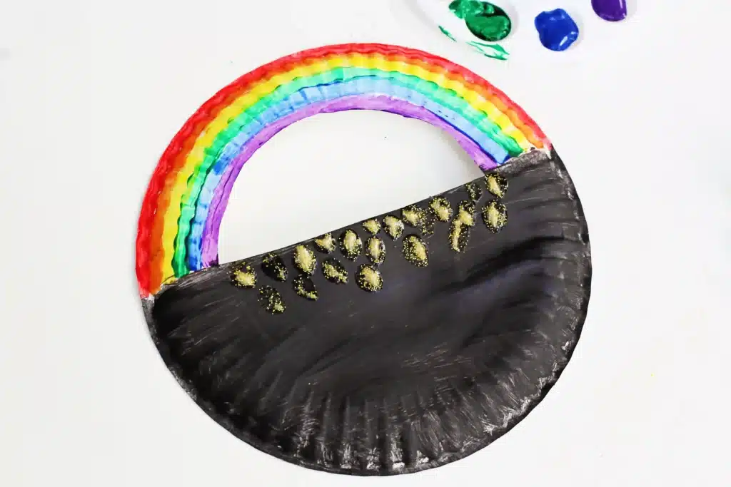 Paper Plate Pot of Gold with Fingerprint Coins - Cultivating Brilliant Minds a paper plate with a semi circle cut out in the middle. the bottom half is painted black with gold fingerprints and the top is painted like a rainbow to make a Paper Plate Pot of Gold with Fingerprint Coins
