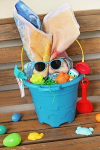 an easter beach bucket with a towel shaped to look like a bunny, sunglasses, flip flops, beach toys, and sunscreen with a printable tag