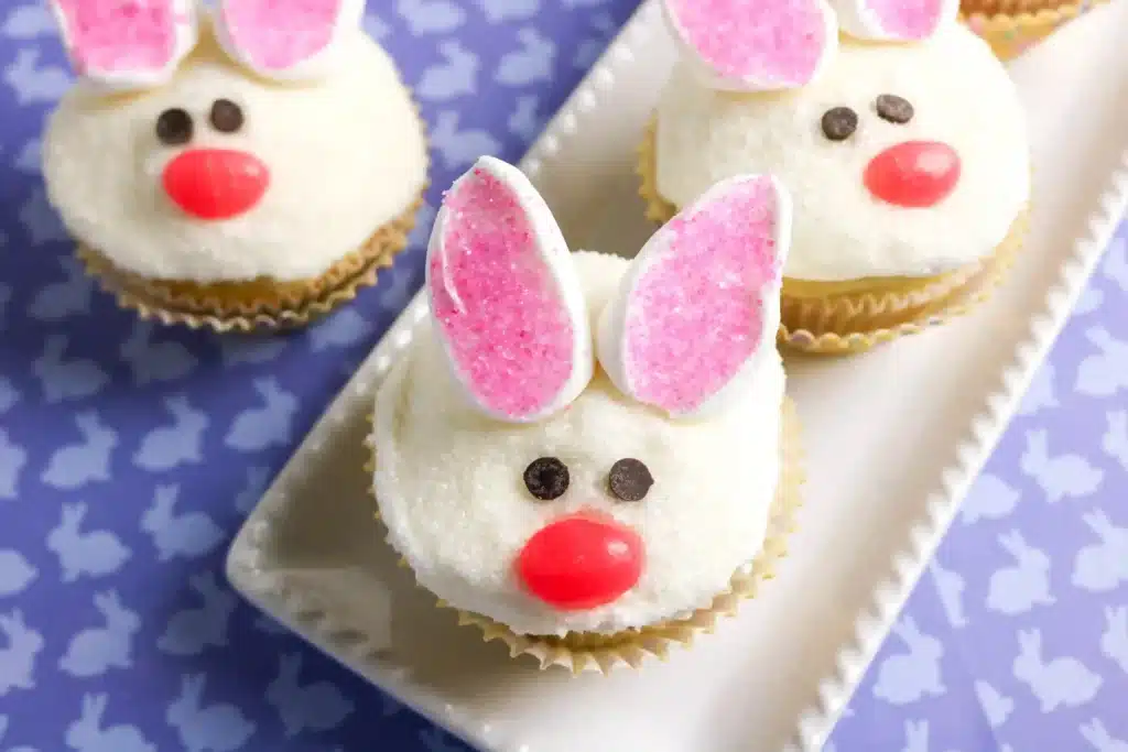 Bunny Cupcakes for Easter - Cultivating Brilliant Minds vanilla cupcakes decorated to look like a bunny with chocolate chip eyes, a jelly bean nose, and cut marshmallows dipped in sanding sugar for ears.