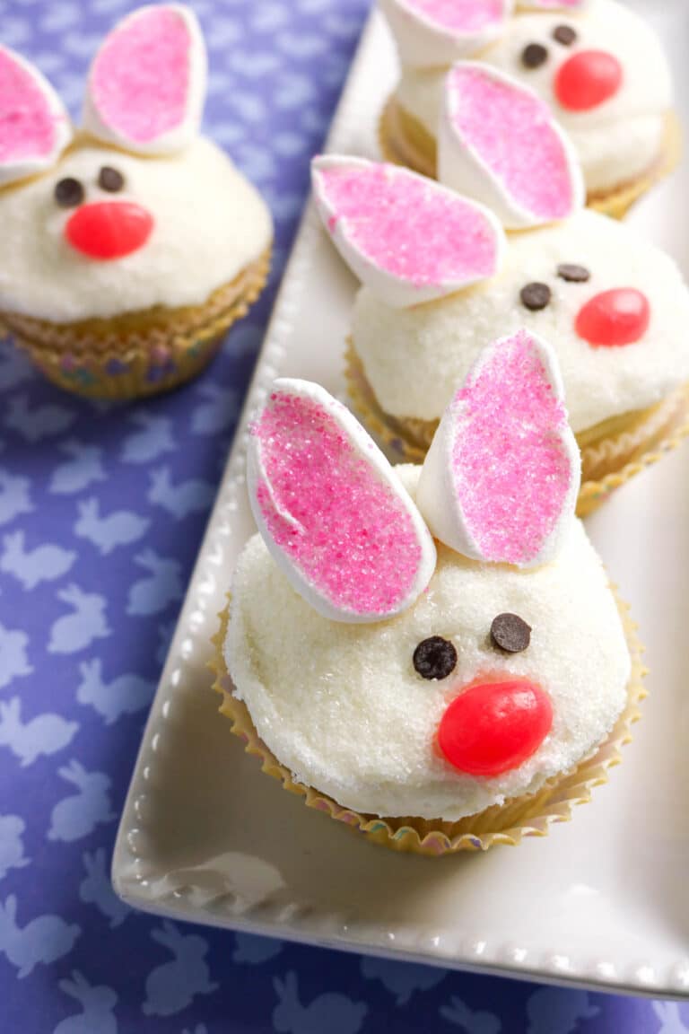 vanilla cupcakes decorated to look like a bunny with chocolate chip eyes, a jelly bean nose, and cut marshmallows dipped in sanding sugar for ears.