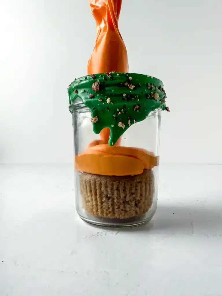 Carrot Cake Jars For Easter - Cultivating Brilliant Minds piping orange buttercream on top of a carrot cupcake inside a jar with green chocolate dripping down the top