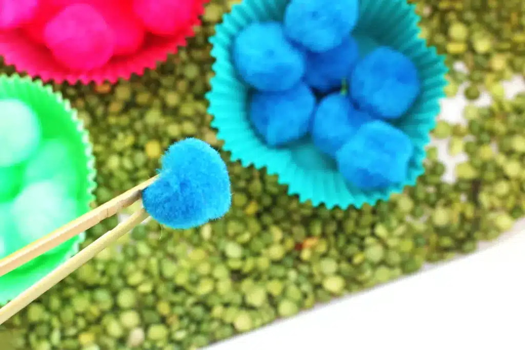 Spring Caterpillar Color Sorting Sensory Bin - Cultivating Brilliant Minds a kid using tongs to pick up blue pom poms